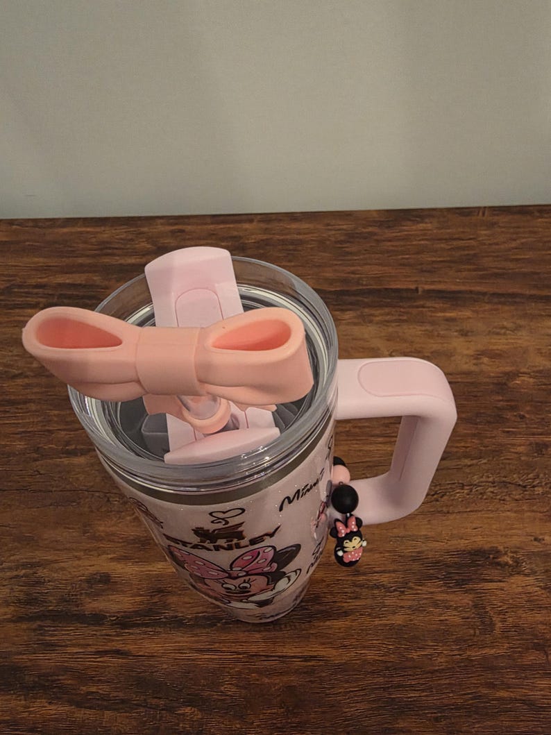 May include: Pink Stanley tumbler with a Minnie Mouse design. The tumbler has a clear lid, a pink handle, and a pink bow-shaped lid closure. A Minnie Mouse charm hangs from the handle. The tumbler is on a wooden surface.