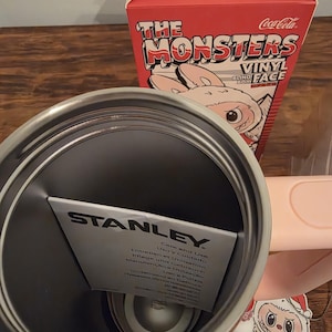 Pu&ograve; includere: Un bicchiere Stanley in acciaio inossidabile con un inserto di carta. Sullo sfondo una scatola rossa con la scritta "The Monsters Vinyl Blind Box". &Egrave; visibile un adesivo Coca-Cola.