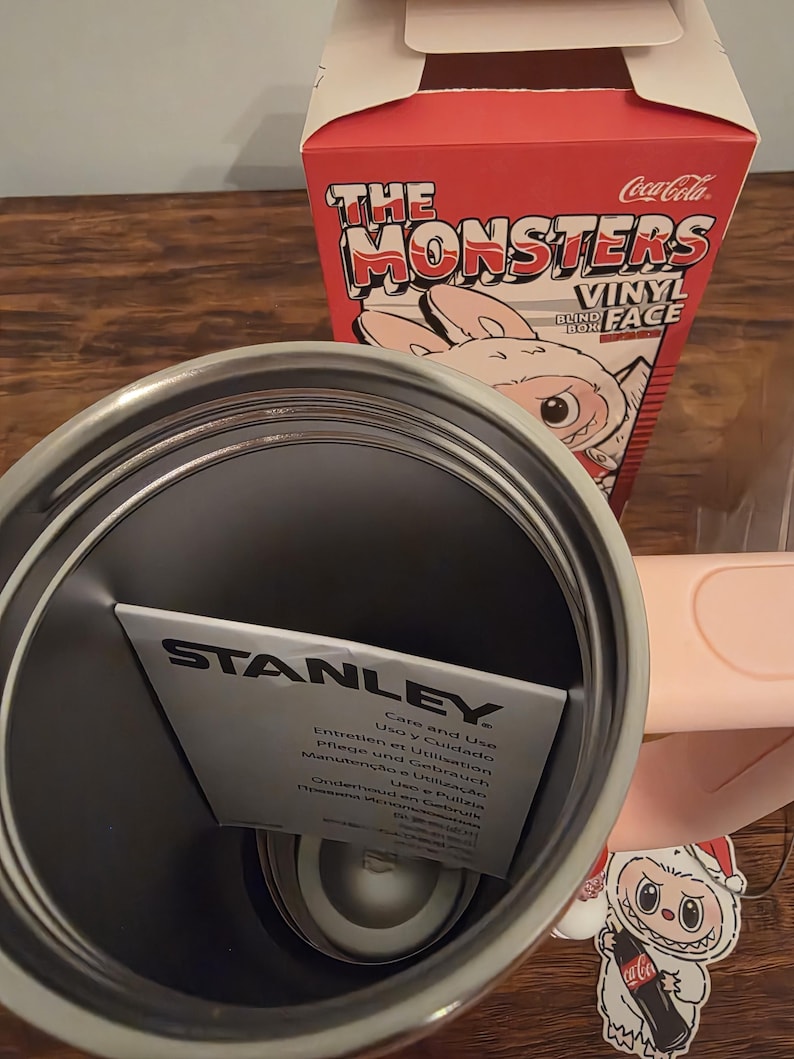 Pu&ograve; includere: Un bicchiere Stanley in acciaio inossidabile con un inserto di carta. Sullo sfondo una scatola rossa con la scritta "The Monsters Vinyl Blind Box". &Egrave; visibile un adesivo Coca-Cola.