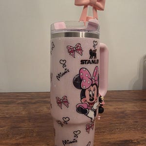 May include: A pink Stanley tumbler with a Minnie Mouse design. The tumbler features a pink bow on the lid and a Minnie Mouse graphic. The tumbler has a handle and is decorated with bows and hearts.