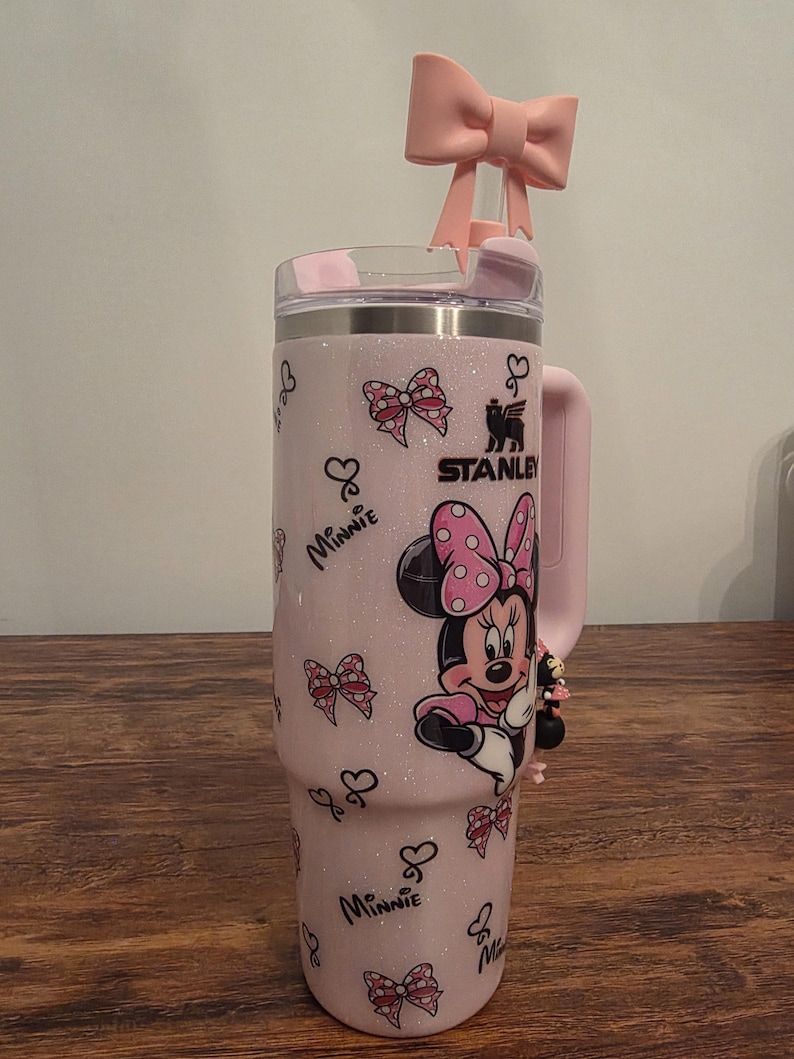 May include: A pink Stanley tumbler with a Minnie Mouse design. The tumbler features a pink bow on the lid and a Minnie Mouse graphic. The tumbler has a handle and is decorated with bows and hearts.