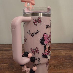 May include: A pink glitter tumbler featuring Minnie Mouse. The tumbler has a pink handle and a pink bow on the lid. The design includes Minnie Mouse, bows, and the word "Minnie". A beaded charm with black and pink beads is attached to the handle.
