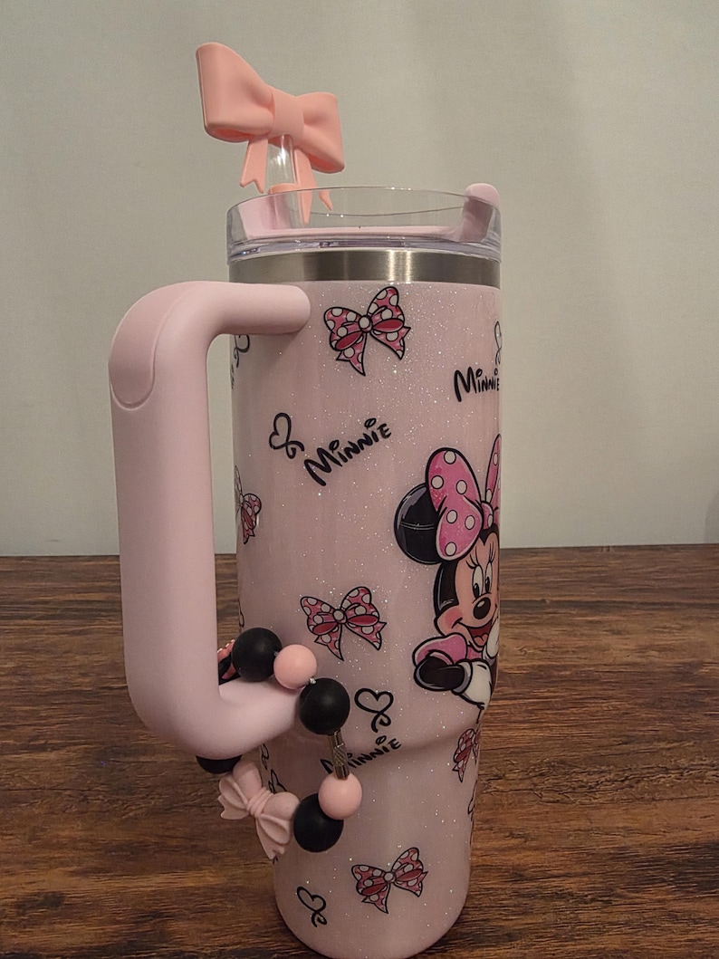 May include: A pink glitter tumbler featuring Minnie Mouse. The tumbler has a pink handle and a pink bow on the lid. The design includes Minnie Mouse, bows, and the word "Minnie". A beaded charm with black and pink beads is attached to the handle.