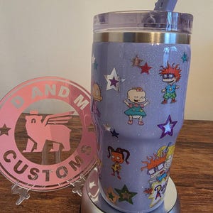 Puede incluir: Un vaso personalizado de color lavanda brillante con personajes de dibujos animados y estrellas. El vaso incluye una tapa transparente y una pajita lavanda a juego. El dise&ntilde;o incorpora personajes de la serie animada Rugrats.