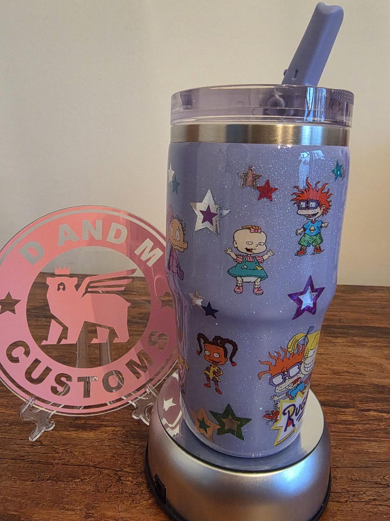 Puede incluir: Un vaso personalizado de color lavanda brillante con personajes de dibujos animados y estrellas. El vaso incluye una tapa transparente y una pajita lavanda a juego. El dise&ntilde;o incorpora personajes de la serie animada Rugrats.