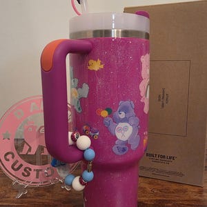 May include: A purple glitter tumbler with a handle and a white lid. The tumbler features Care Bears graphics and a beaded charm. It has a straw and a handle with an orange accent. The text "BUILT FOR LIFE" is visible.