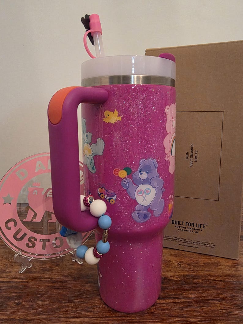 May include: A purple glitter tumbler with a handle and a white lid. The tumbler features Care Bears graphics and a beaded charm. It has a straw and a handle with an orange accent. The text "BUILT FOR LIFE" is visible.