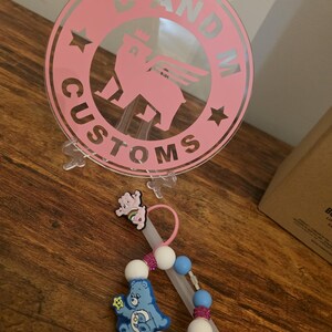 May include: A pink circular logo with the text "D AND M CUSTOMS" and a bear graphic. A pacifier clip with blue and white beads, a blue Care Bear, and a pink Care Bear are also visible.
