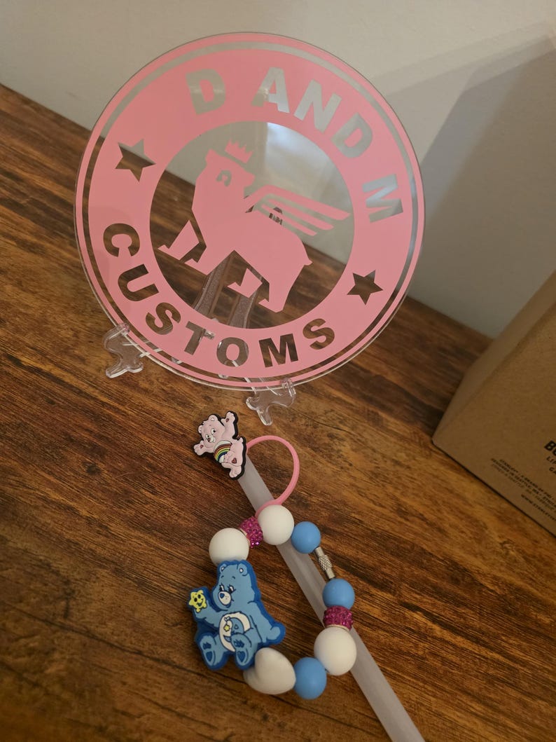 May include: A pink circular logo with the text "D AND M CUSTOMS" and a bear graphic. A pacifier clip with blue and white beads, a blue Care Bear, and a pink Care Bear are also visible.