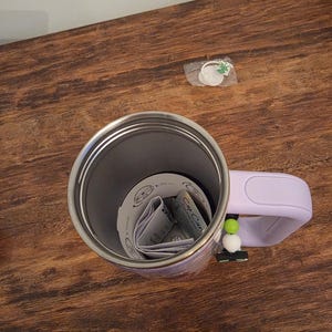 May include: A stainless steel travel mug with a light purple handle and a decorative charm. The mug contains folded paper inserts. The charm has green and white beads. The mug sits on a wooden surface.