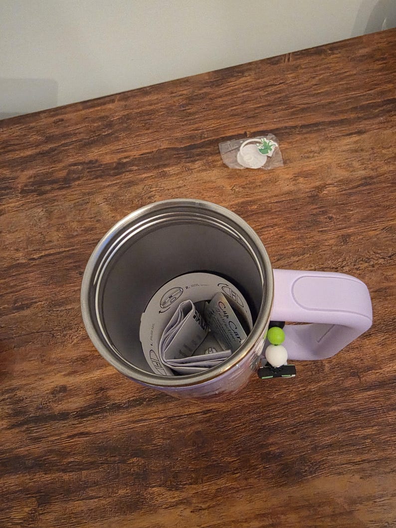May include: A stainless steel travel mug with a light purple handle and a decorative charm. The mug contains folded paper inserts. The charm has green and white beads. The mug sits on a wooden surface.