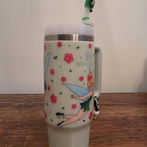 Puede incluir: Un vaso Stanley verde claro con tapa y asa blancas. El vaso presenta un dise&ntilde;o de hada con flores rosas y corazones verdes. Una pajita verde y blanca con un adorno de hada est&aacute; insertada en la tapa.