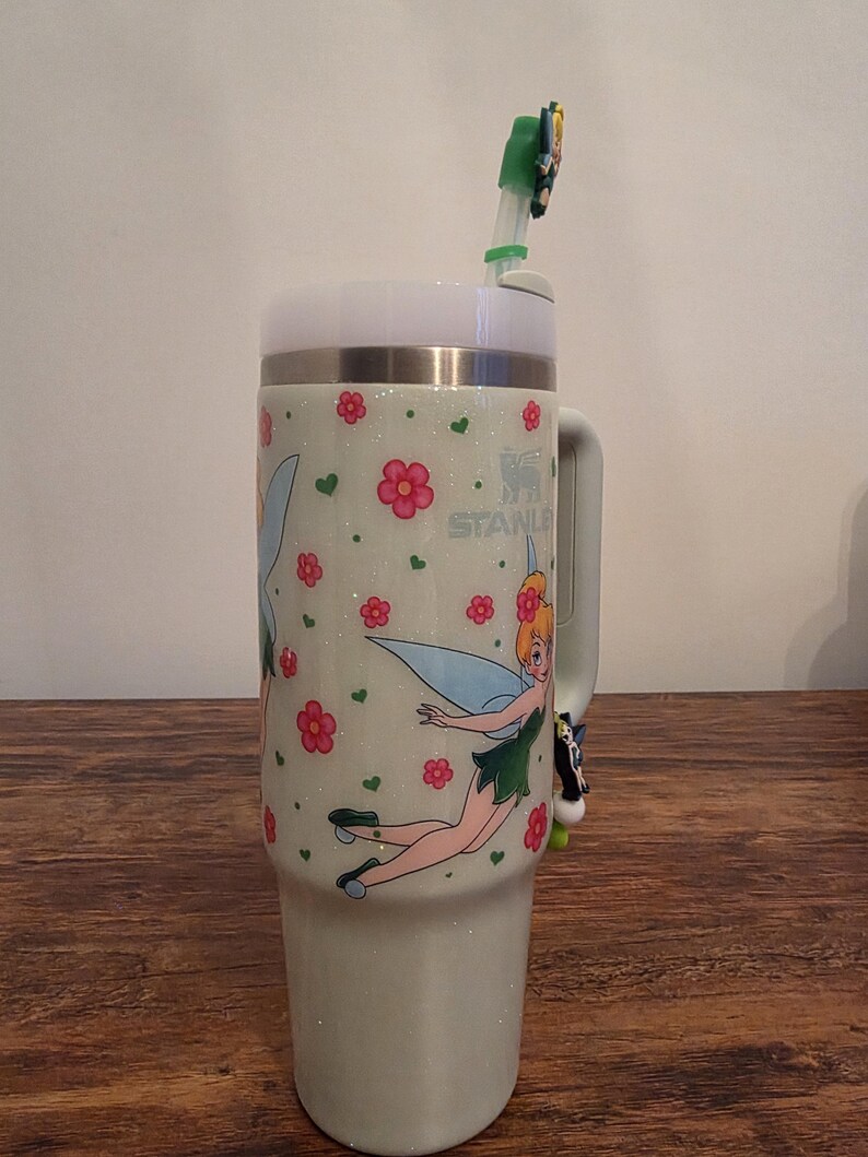 Puede incluir: Un vaso Stanley verde claro con tapa y asa blancas. El vaso presenta un dise&ntilde;o de hada con flores rosas y corazones verdes. Una pajita verde y blanca con un adorno de hada est&aacute; insertada en la tapa.