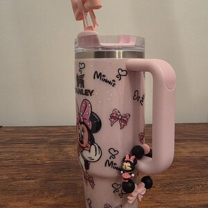 May include: A pink insulated tumbler featuring Minnie Mouse graphics and a matching handle. The tumbler has a bow-shaped straw topper and a Minnie Mouse charm. The tumbler is approximately 10 inches tall.