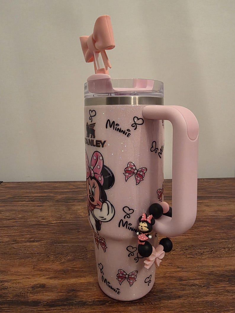 May include: A pink insulated tumbler featuring Minnie Mouse graphics and a matching handle. The tumbler has a bow-shaped straw topper and a Minnie Mouse charm. The tumbler is approximately 10 inches tall.