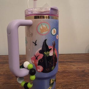 May include: A stainless steel tumbler with a purple handle and lid, featuring a design with a witch illustration and other graphics. The tumbler has a beaded charm bracelet attached to the handle. The tumbler is decorated with a gradient of pink, blue, and purple.