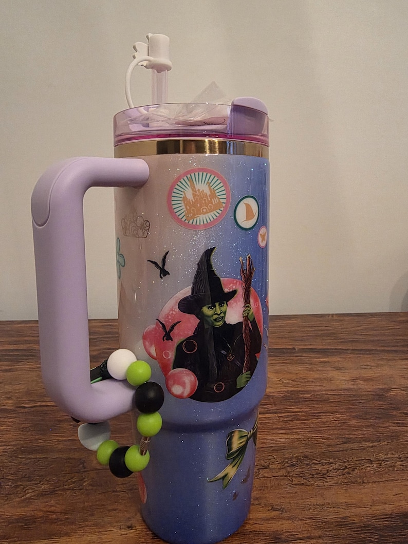 May include: A stainless steel tumbler with a purple handle and lid, featuring a design with a witch illustration and other graphics. The tumbler has a beaded charm bracelet attached to the handle. The tumbler is decorated with a gradient of pink, blue, and purple.