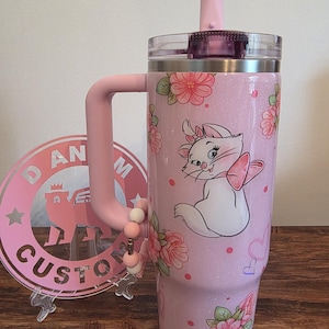 May include: A light pink glitter tumbler with a handle, a clear lid, and a straw. The tumbler features a graphic of a white kitten with a pink bow, surrounded by pink floral patterns, small pink dots, and heart outlines. A beaded charm hangs from the handle.