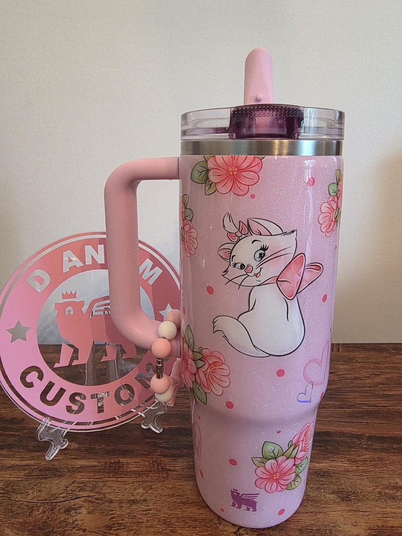 May include: A light pink glitter tumbler with a handle, a clear lid, and a straw. The tumbler features a graphic of a white kitten with a pink bow, surrounded by pink floral patterns, small pink dots, and heart outlines. A beaded charm hangs from the handle.