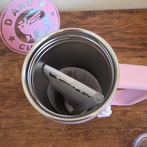 May include: A light pink Stanley tumbler with a handle, shown from an overhead perspective. Inside the metal interior sits a white paper instruction manual and a gray straw. A circular pink lid with a winged lion logo and the text D AND M CU is nearby.