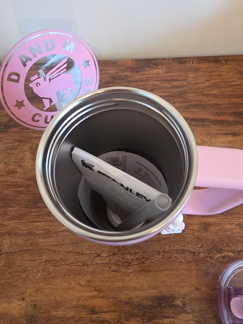 May include: A light pink Stanley tumbler with a handle, shown from an overhead perspective. Inside the metal interior sits a white paper instruction manual and a gray straw. A circular pink lid with a winged lion logo and the text D AND M CU is nearby.