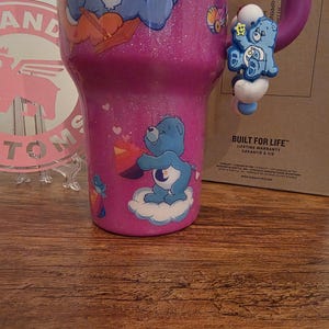 May include: A pink glitter tumbler featuring Care Bears characters. The tumbler has a blue Care Bear charm attached to the handle. The tumbler is decorated with various Care Bear graphics and cloud designs. A pink and white logo is visible in the background.