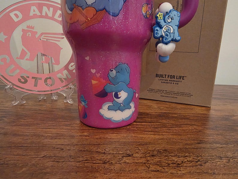 May include: A pink glitter tumbler featuring Care Bears characters. The tumbler has a blue Care Bear charm attached to the handle. The tumbler is decorated with various Care Bear graphics and cloud designs. A pink and white logo is visible in the background.
