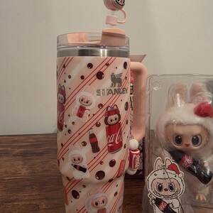 Pu&ograve; includere: Un bicchiere Stanley rosa e bianco con un design a coniglietto e Coca-Cola. Il bicchiere ha un manico rosa abbinato e un tappo per cannuccia a forma di coniglietto. Una statuina di coniglietto &egrave; visibile sullo sfondo.