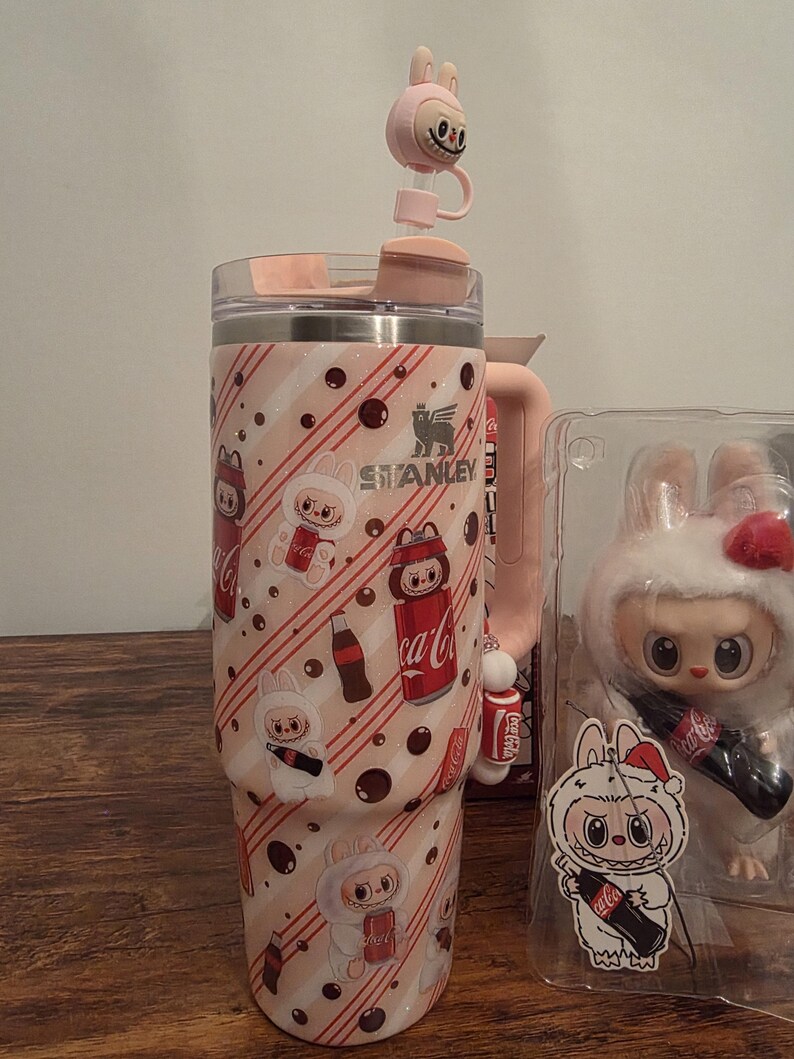 Pu&ograve; includere: Un bicchiere Stanley rosa e bianco con un design a coniglietto e Coca-Cola. Il bicchiere ha un manico rosa abbinato e un tappo per cannuccia a forma di coniglietto. Una statuina di coniglietto &egrave; visibile sullo sfondo.