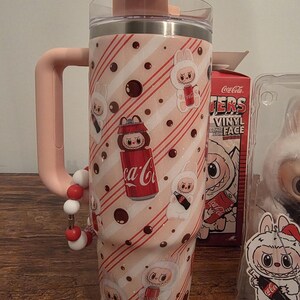 Pu&ograve; includere: Un bicchiere rosa e bianco con manico e coperchio a forma di coniglietto. Il bicchiere presenta un motivo ripetuto di personaggi di coniglietti, lattine di Coca-Cola e strisce diagonali. Un cinturino da polso con perline &egrave; attaccato al manico. Una scatola di Coca-Cola &egrave; visibile sullo sfondo.