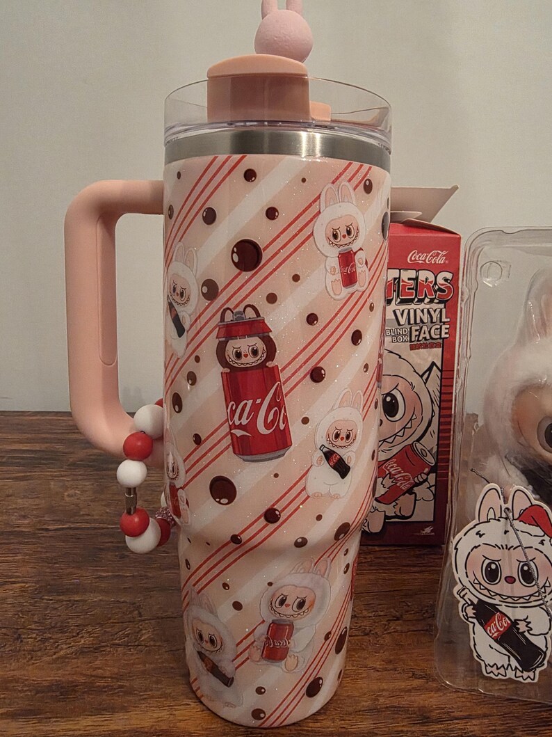Pu&ograve; includere: Un bicchiere rosa e bianco con manico e coperchio a forma di coniglietto. Il bicchiere presenta un motivo ripetuto di personaggi di coniglietti, lattine di Coca-Cola e strisce diagonali. Un cinturino da polso con perline &egrave; attaccato al manico. Una scatola di Coca-Cola &egrave; visibile sullo sfondo.