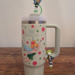 Puede incluir: Un vaso Stanley verde claro con asa, decorado con una imagen de dibujos animados de Campanilla y flores rosas. El vaso tiene una tapa blanca y una pajita verde con un adorno de Campanilla. La palabra "STANLEY" est&aacute; impresa en el vaso.