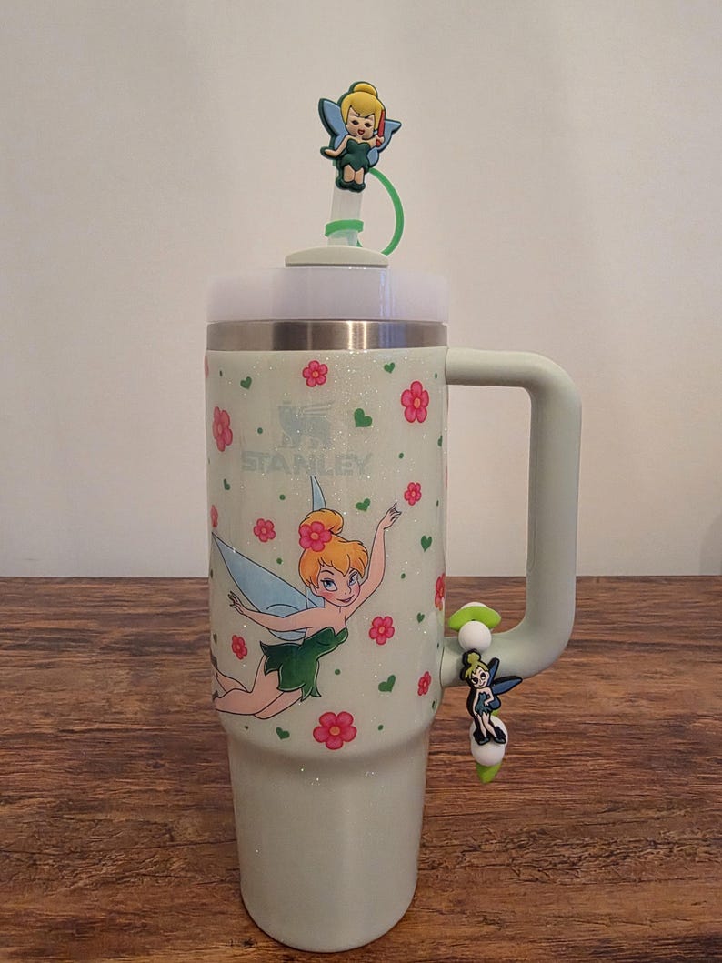 Puede incluir: Un vaso Stanley verde claro con asa, decorado con una imagen de dibujos animados de Campanilla y flores rosas. El vaso tiene una tapa blanca y una pajita verde con un adorno de Campanilla. La palabra "STANLEY" est&aacute; impresa en el vaso.