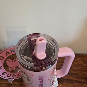 May include: A light pink Stanley tumbler featuring a Marie cat graphic, a clear lid with a pink straw opening, and a matching pink handle. A small beaded charm with a cat figure hangs from the handle, and the Stanley logo is printed on the side.