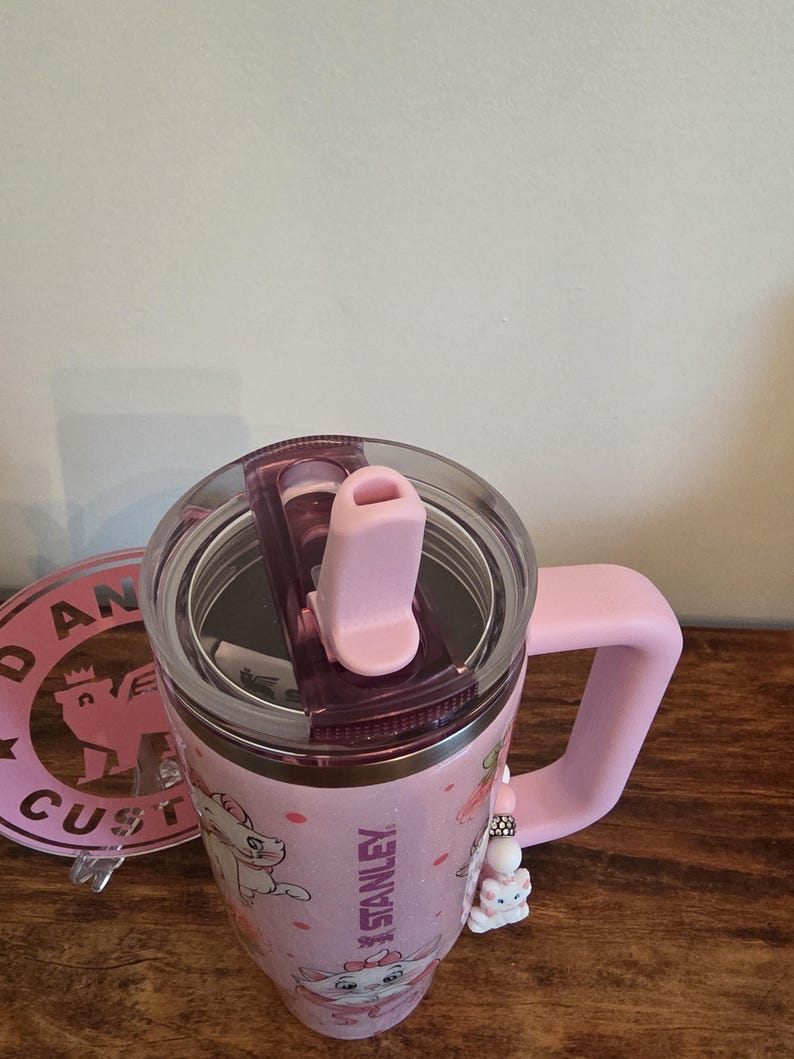 May include: A light pink Stanley tumbler featuring a Marie cat graphic, a clear lid with a pink straw opening, and a matching pink handle. A small beaded charm with a cat figure hangs from the handle, and the Stanley logo is printed on the side.