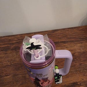 May include: A lavender Stanley tumbler with a handle, featuring a clear lid and a black witch-themed charm. The tumbler has a graphic of characters from the musical "Wicked" and a matching charm.