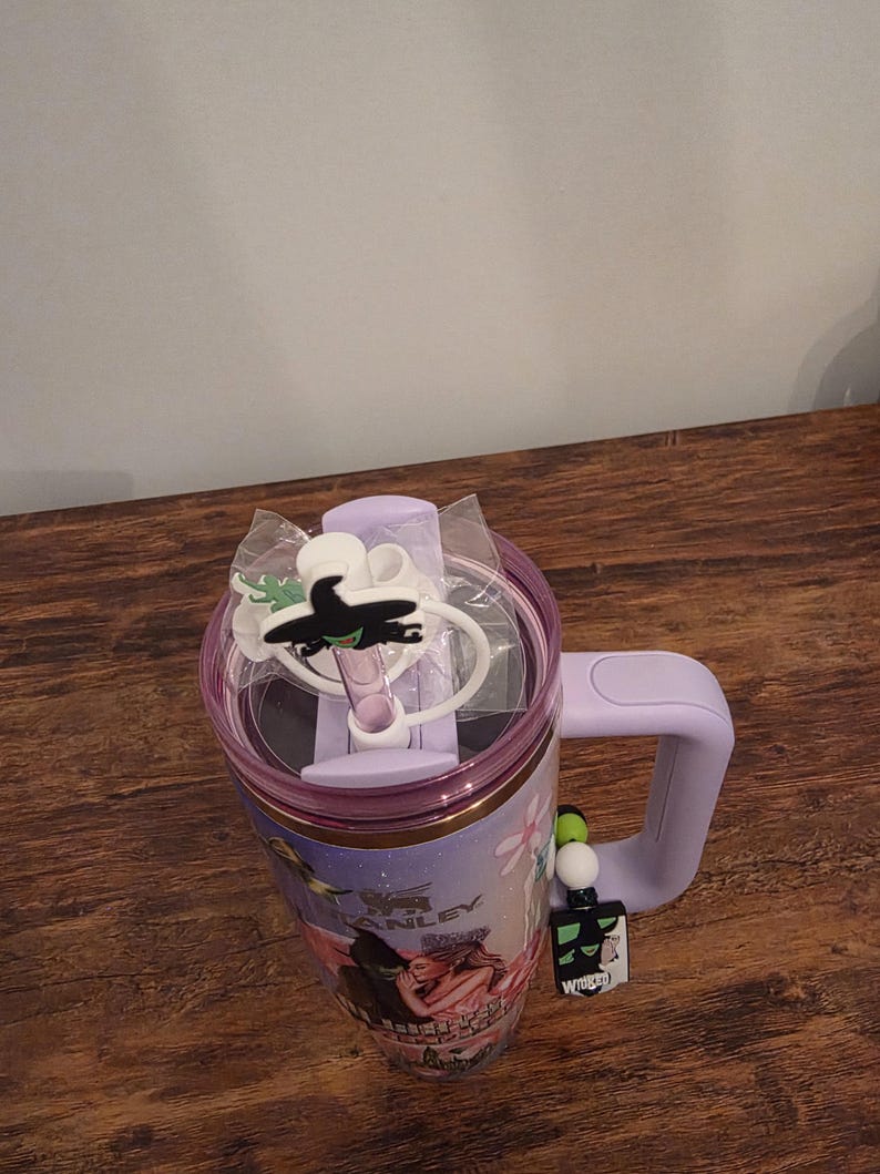 May include: A lavender Stanley tumbler with a handle, featuring a clear lid and a black witch-themed charm. The tumbler has a graphic of characters from the musical "Wicked" and a matching charm.