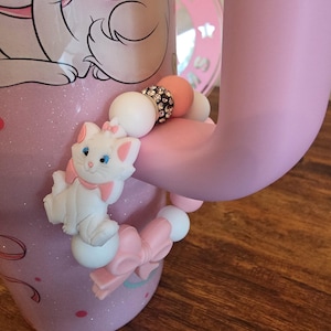 May include: A beaded accessory attached to a light pink mug handle, featuring a white cat character with a pink bow, round white and pink silicone beads, and a single bead encrusted with small clear crystals, all arranged in a circular pattern.