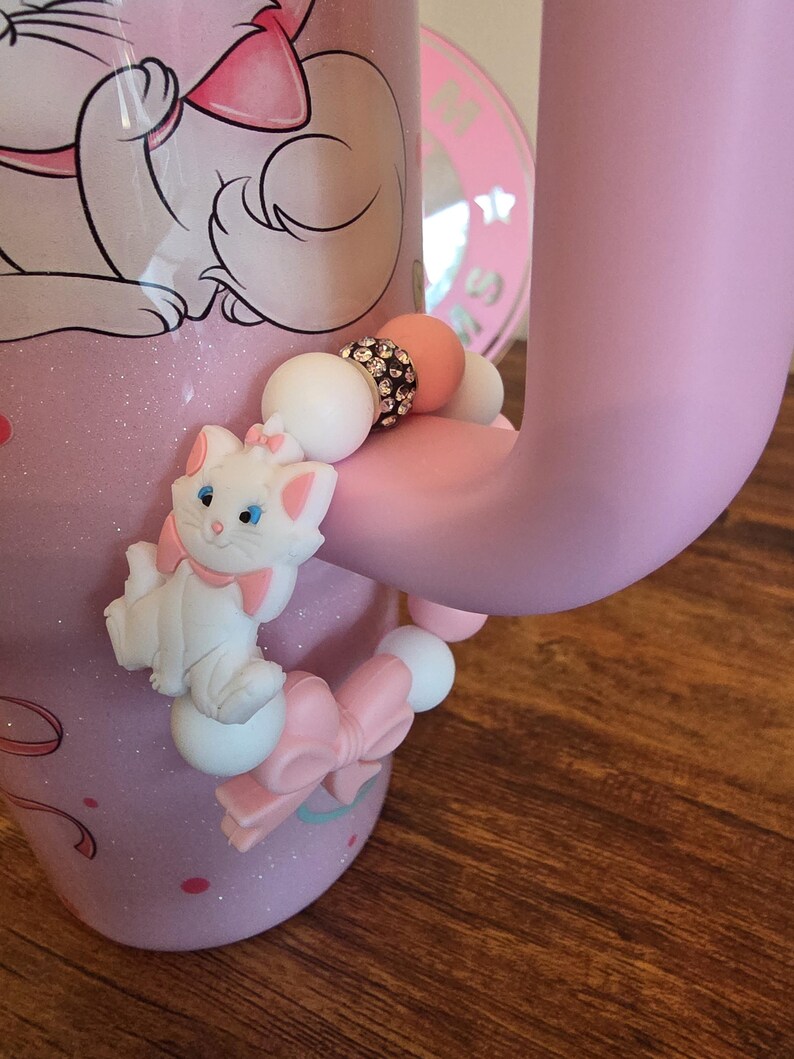 May include: A beaded accessory attached to a light pink mug handle, featuring a white cat character with a pink bow, round white and pink silicone beads, and a single bead encrusted with small clear crystals, all arranged in a circular pattern.
