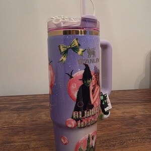 May include: A purple Stanley tumbler with a handle, featuring a design of a witch and a woman. The tumbler has a straw and a decorative witch hat on top. The design includes a green bow and the Stanley logo.