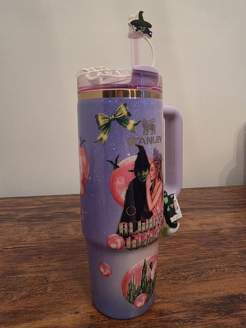 May include: A purple Stanley tumbler with a handle, featuring a design of a witch and a woman. The tumbler has a straw and a decorative witch hat on top. The design includes a green bow and the Stanley logo.
