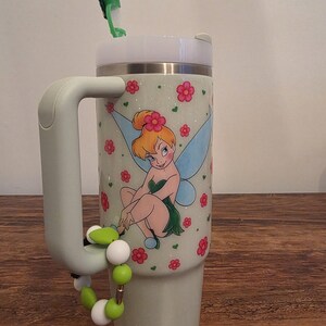 Puede incluir: Un vaso de color verde claro con tapa y asa blancas. El vaso presenta una imagen de dibujos animados de Campanilla rodeada de flores rosas. Un accesorio de cuentas verdes y blancas est&aacute; sujeto al asa. Una pajita verde est&aacute; insertada en la tapa.