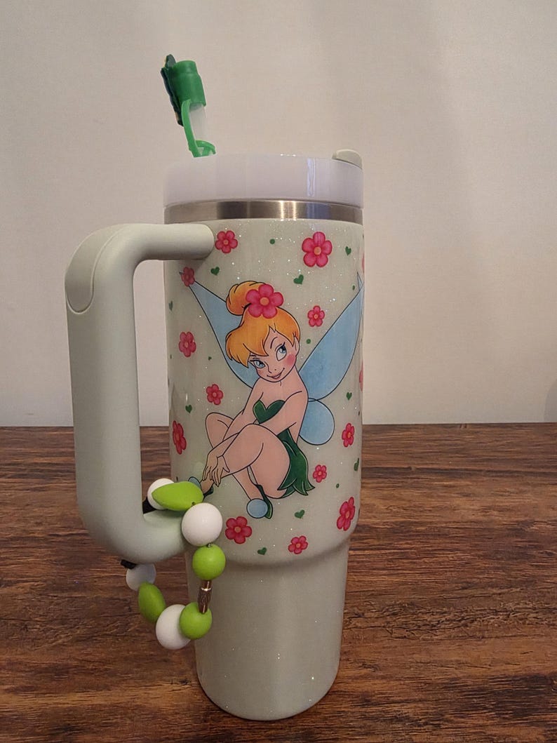 Puede incluir: Un vaso de color verde claro con tapa y asa blancas. El vaso presenta una imagen de dibujos animados de Campanilla rodeada de flores rosas. Un accesorio de cuentas verdes y blancas est&aacute; sujeto al asa. Una pajita verde est&aacute; insertada en la tapa.