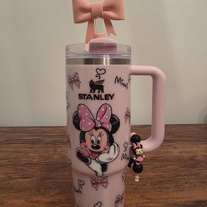 May include: A pink Stanley tumbler featuring Minnie Mouse. The tumbler has a pink handle, a bow on the lid, and a charm. The design includes Minnie Mouse, bows, and the word "Minnie".