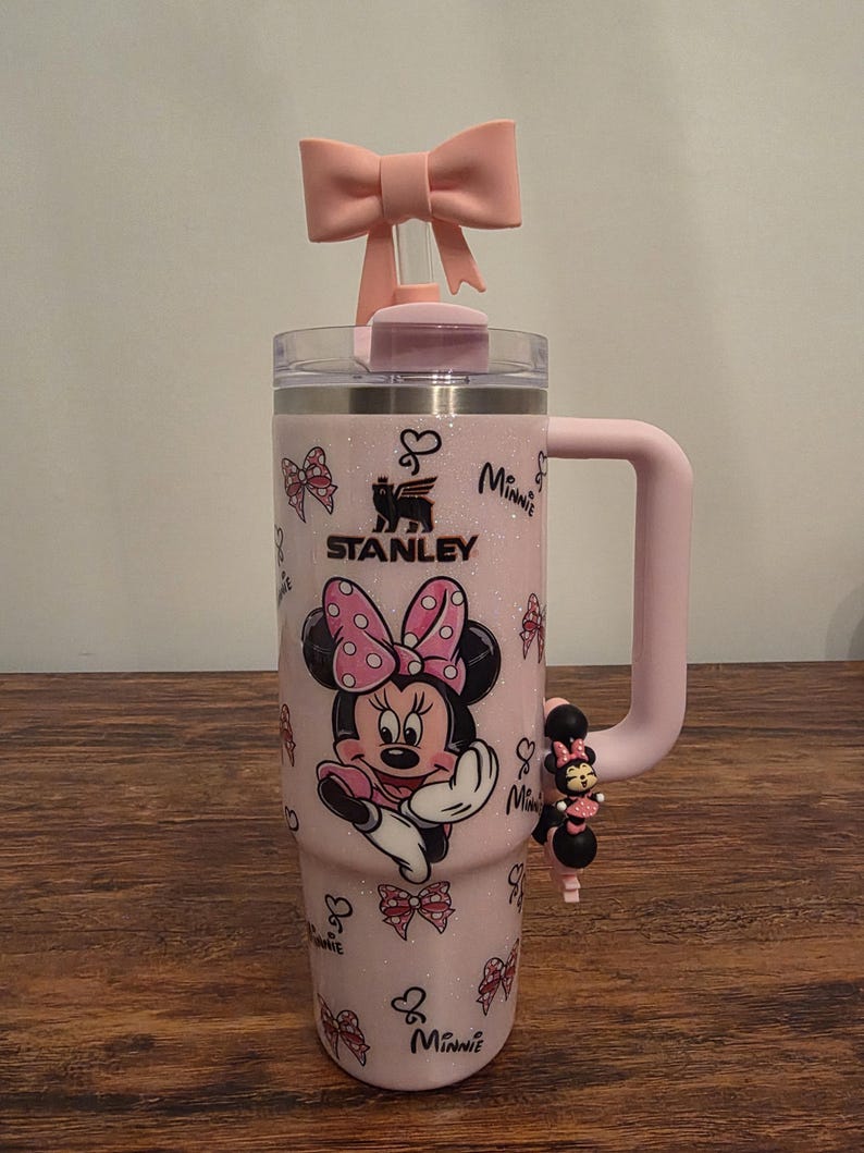May include: A pink Stanley tumbler featuring Minnie Mouse. The tumbler has a pink handle, a bow on the lid, and a charm. The design includes Minnie Mouse, bows, and the word "Minnie".