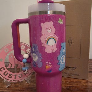 May include: A glittery magenta tumbler with a handle, featuring Care Bear graphics. The tumbler has a white top, a pink lid with a straw, and a beaded charm. The design includes a rainbow and various Care Bears.