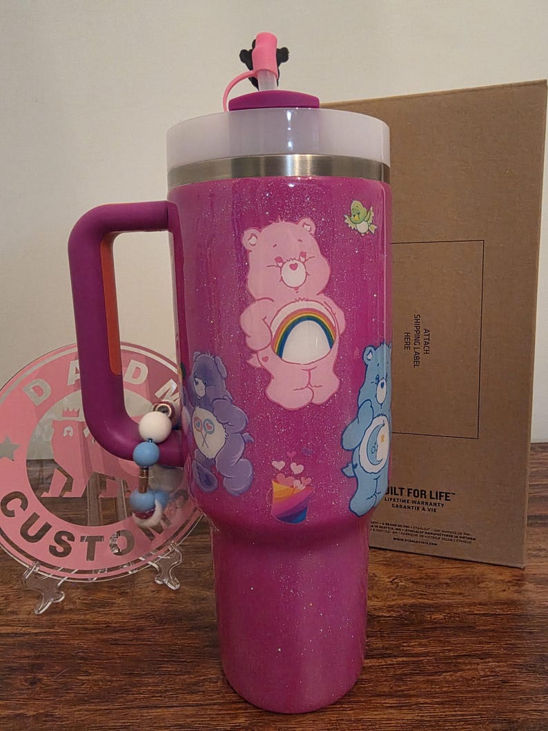 May include: A glittery magenta tumbler with a handle, featuring Care Bear graphics. The tumbler has a white top, a pink lid with a straw, and a beaded charm. The design includes a rainbow and various Care Bears.