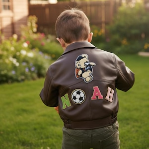 May include: A brown leather jacket with a cartoon patch on the back, spelling the name "NOAH" with a soccer ball graphic. The jacket is worn by a child in a garden with green grass and a wooden fence.