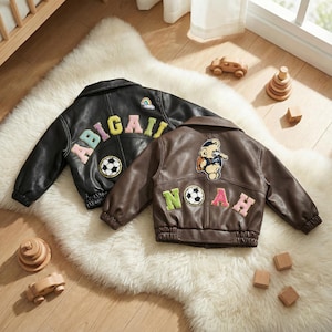 Op de afbeelding: Twee leren kinderjassen, een zwarte en een bruine, liggen op een witte, pluizige vloerkleed. De zwarte jas heeft de naam "ABIGAIL" en een voetbalpatch. De bruine jas heeft de naam "NOAH" en een teddybeerpatch.