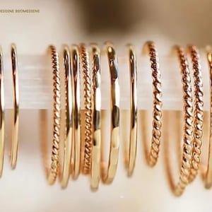 Può includere: Una collezione di anelli sottili in oro disposti in fila su una barra orizzontale. Il set include varie texture, con fasce lisce, design a corda intrecciata e finiture martellate, tutti caratterizzati da una superficie dorata lucida.